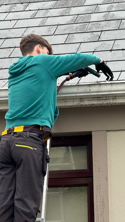 Worker in teal hoodie and safety harness cleaning or inspecting gray roof shingles above a window