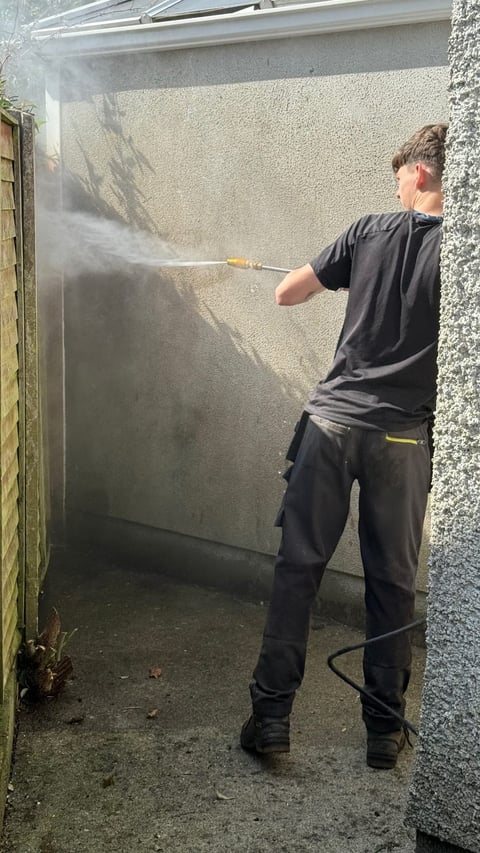 Worker pressure washing a concrete wall in a narrow alley using a high-pressure spray nozzle
