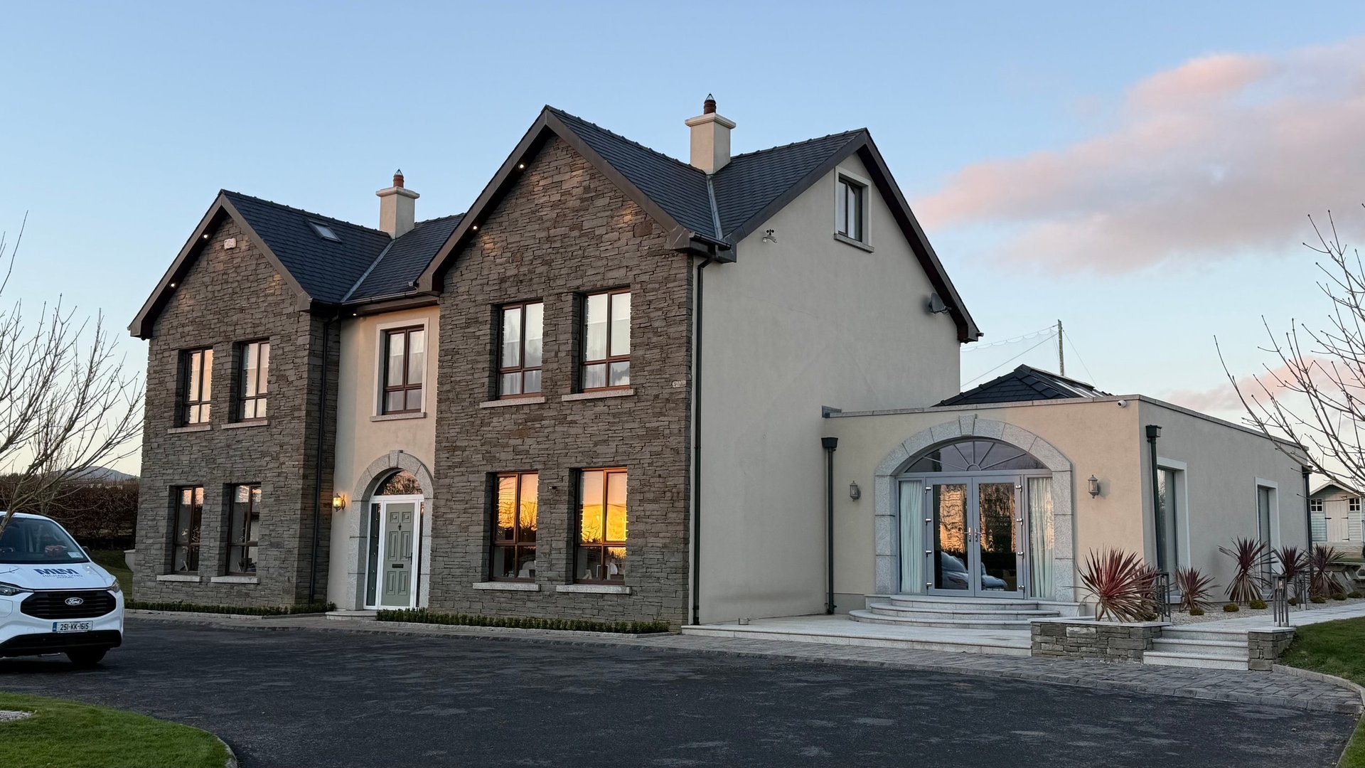 Modern two-story stone and rendered house with illuminated windows, driveway, and contemporary extension at dusk