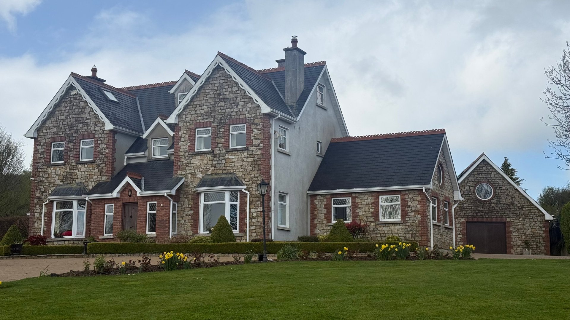 Large brick and stone two-story house with slate roof, multiple chimneys, manicured lawn, and landscaping shrubs on a clear day