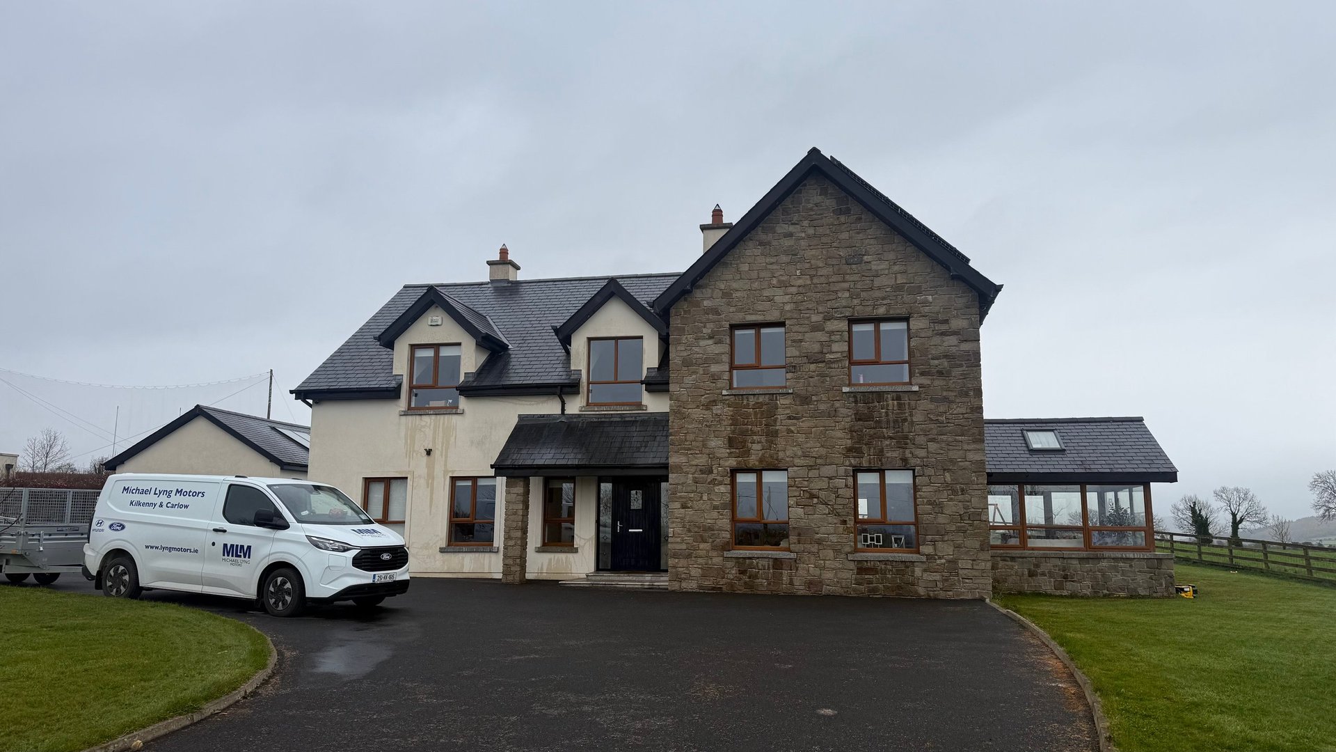 Two-story stone residential house with a cream-colored addition and a company van parked in the driveway, set in rural countryside