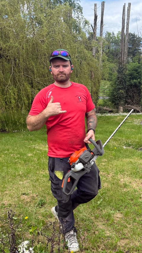 Man in red shirt and blue cap holding a chainsaw in a grassy backyard with trees in background