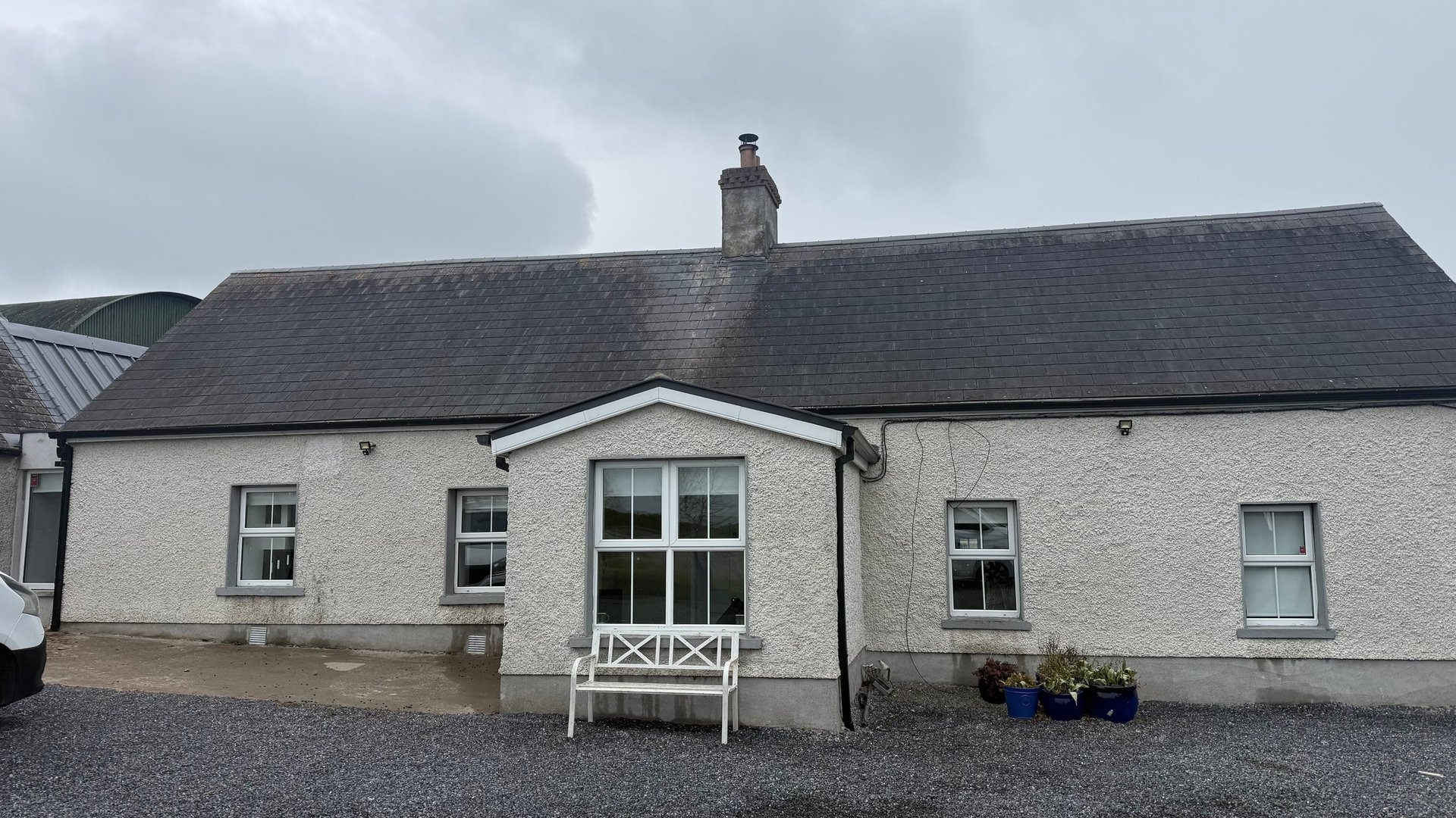 White-walled rural cottage with dark slate roof, central entrance with white railing, and gravel driveway