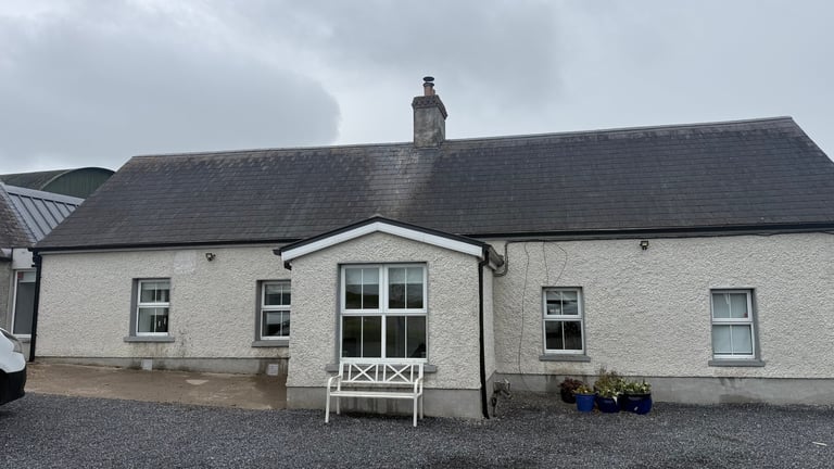 White-walled rural cottage with dark slate roof, central entrance with white railing, and gravel driveway on an overcast day